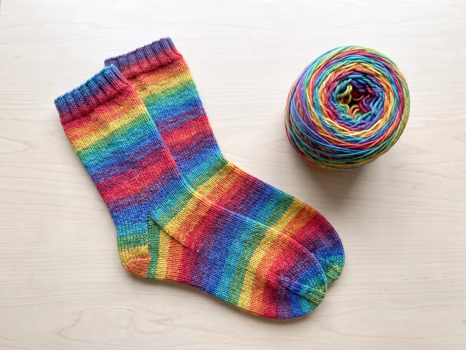 Handgestrickte Socken aus Farbverlaufsgarn mit bunten Streifen neben einem Wollknäuel.